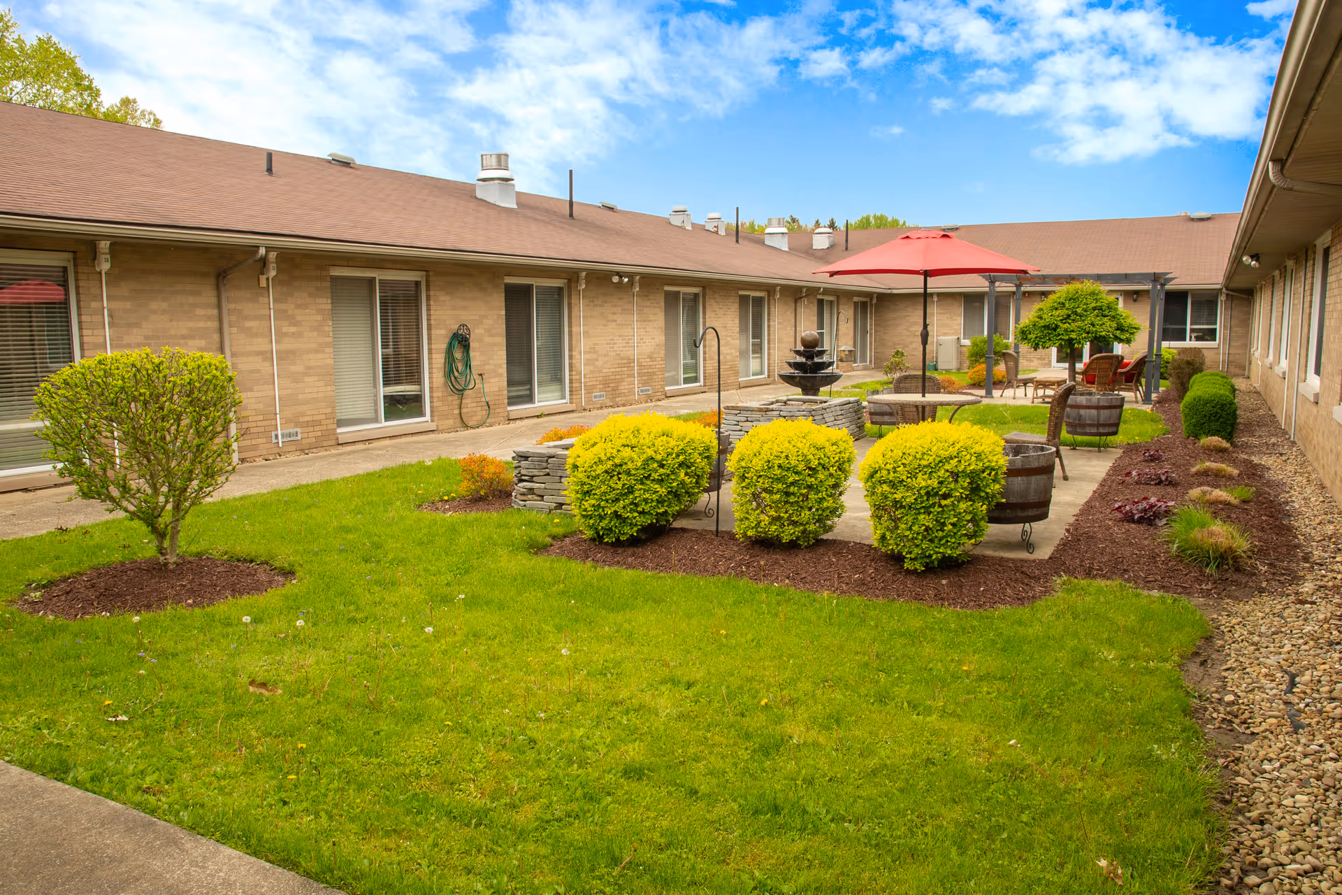 A courtyard area in a senior living facility with green grass, neatly trimmed bushes, a stone water fountain, patio tables with red umbrellas, and chairs. The courtyard is surrounded by a single-story brick building with windows and sliding glass doors.