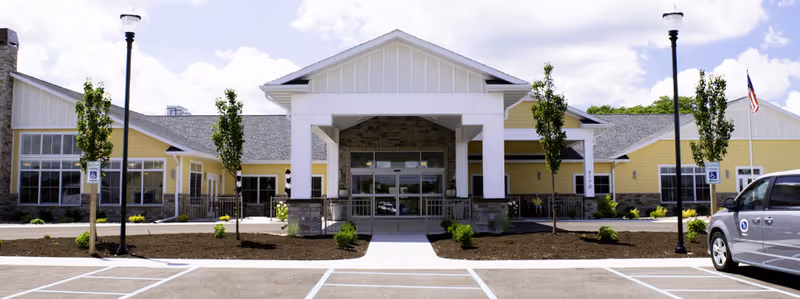 Front entrance of a single-story yellow assisted living building with a covered porte-cochère, parking spaces, lamp posts, and small trees.