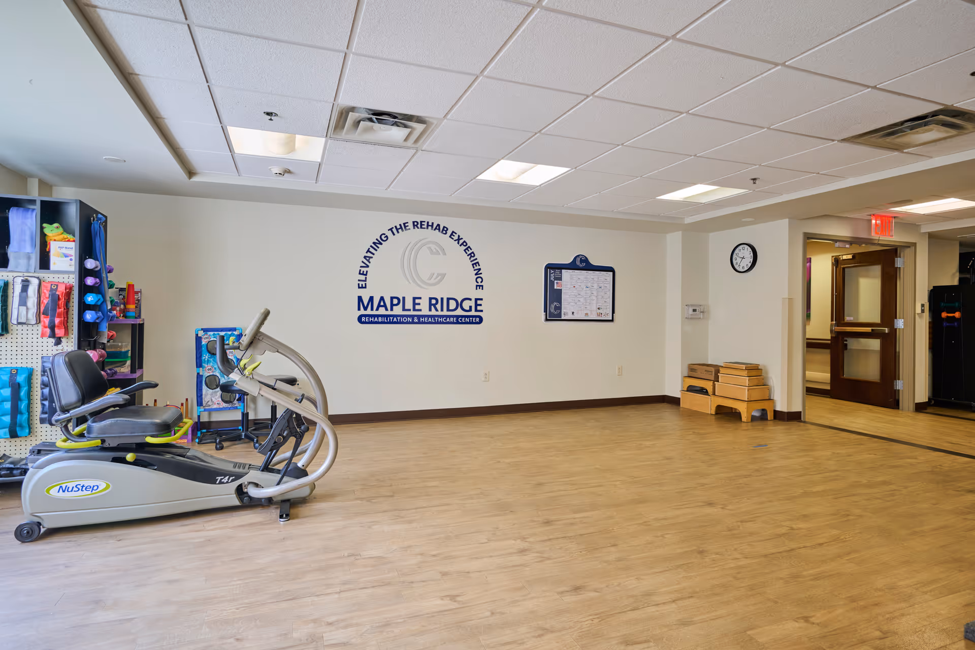 A rehabilitation exercise room with a NuStep exercise machine, various therapy equipment on shelves, a clock on the wall, and a sign that reads 'Elevating the Rehab Experience Maple Ridge Rehabilitation & Healthcare Center'. The room has wood flooring and a drop ceiling with fluorescent lights.