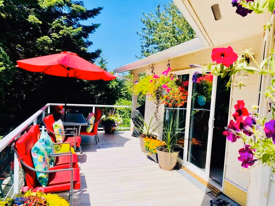 Outdoor patio area with red cushioned chairs and colorful floral pillows, a table with a red umbrella, hanging flower baskets, potted plants, and a sliding glass door leading inside. The patio is surrounded by green trees under a clear blue sky.
