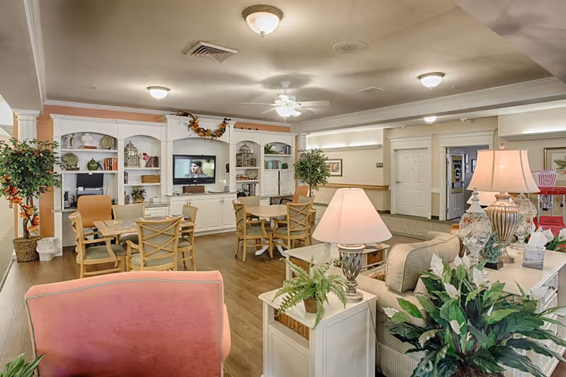 A cozy senior living common area with wooden floors, several tables and chairs, a large white entertainment center with a TV, decorative plants, and warm lighting from ceiling fixtures and table lamps.
