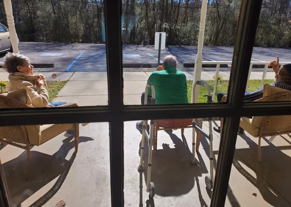 View through a window showing three elderly people sitting outside on a porch. Two are seated in chairs and one is seated on a walker. They are enjoying the sunny day with a parking lot and trees in the background.