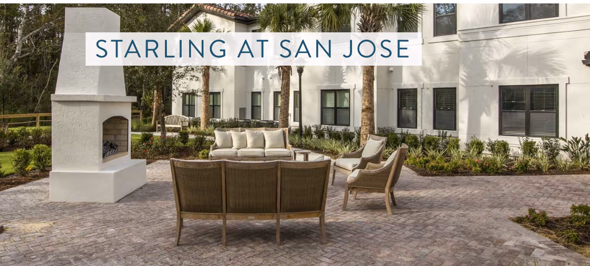 Outdoor patio area at Starling at San Jose featuring a white outdoor fireplace, cushioned sofa, and several wicker chairs arranged on a brick-paved surface with a building and palm trees in the background.