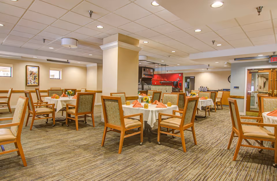 Well-lit dining room with round tables set with white tablecloths, napkins and wooden chairs.