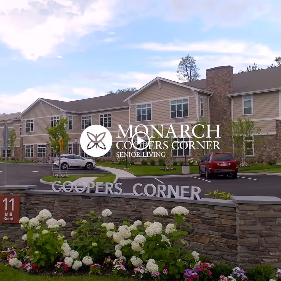 Exterior view of Monarch Coopers Corner senior living facility showing a two-story building with beige siding and stone accents, a parking lot with cars, a landscaped area with flowers and bushes, and a stone sign that reads 'COOPERS CORNER' with the address '11 Mill Road'.