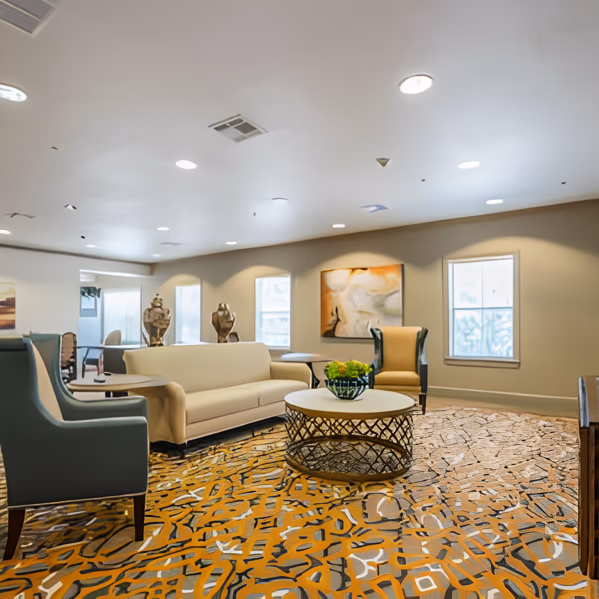 A spacious living room with a beige sofa, two armchairs, a round coffee table with a small plant, abstract wall art, and large windows allowing natural light to fill the room. The carpet has an intricate pattern in shades of orange, brown, and beige.