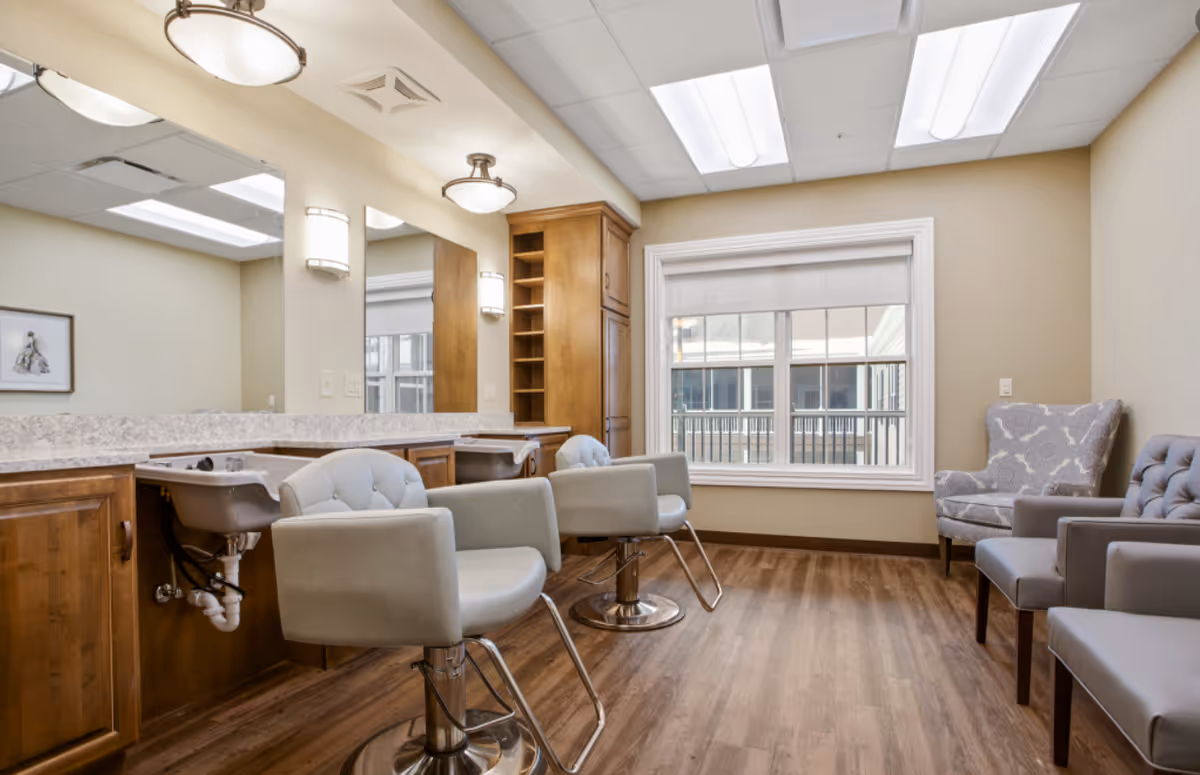A bright and clean salon area with two light gray salon chairs in front of a long mirror and countertop with built-in sinks. There are wooden cabinets and shelves along the wall, a large window with a white blind, and additional seating with two upholstered armchairs on the right side. The room has wood flooring and soft beige walls.