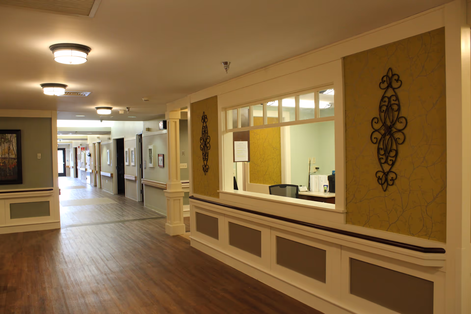 Interior hallway of a senior living facility with wood flooring, beige walls, and ceiling lights. There is a reception window with a desk and chair behind it on the right side, decorative wall art, and framed pictures along the hallway walls.