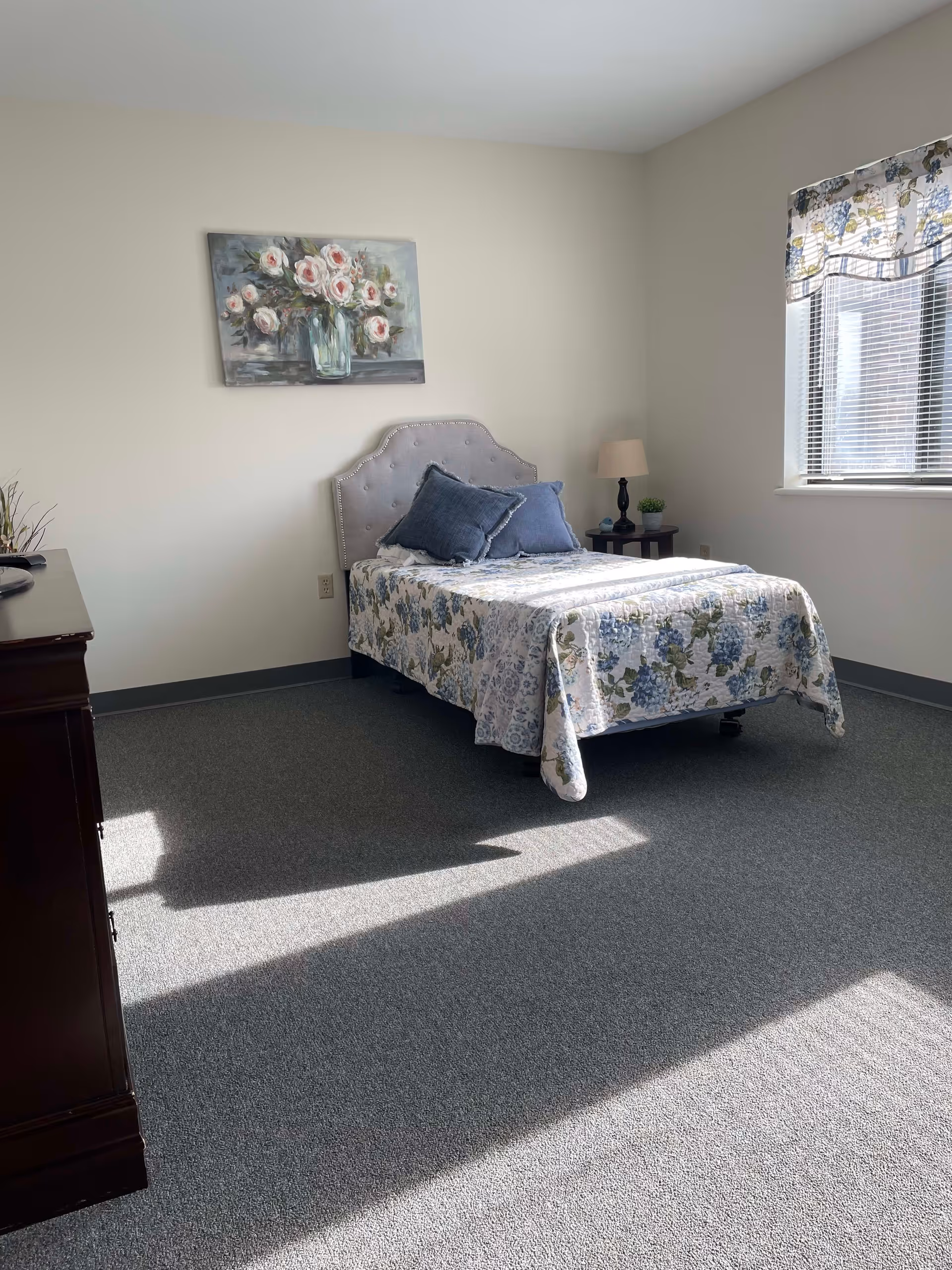 A bright bedroom with a single bed covered in a floral quilt and two blue pillows. The bed has a tufted headboard. There is a nightstand with a lamp and a small plant next to the bed. A window with floral curtains lets in sunlight, casting shadows on the carpeted floor. A floral painting hangs on the wall above the bed.
