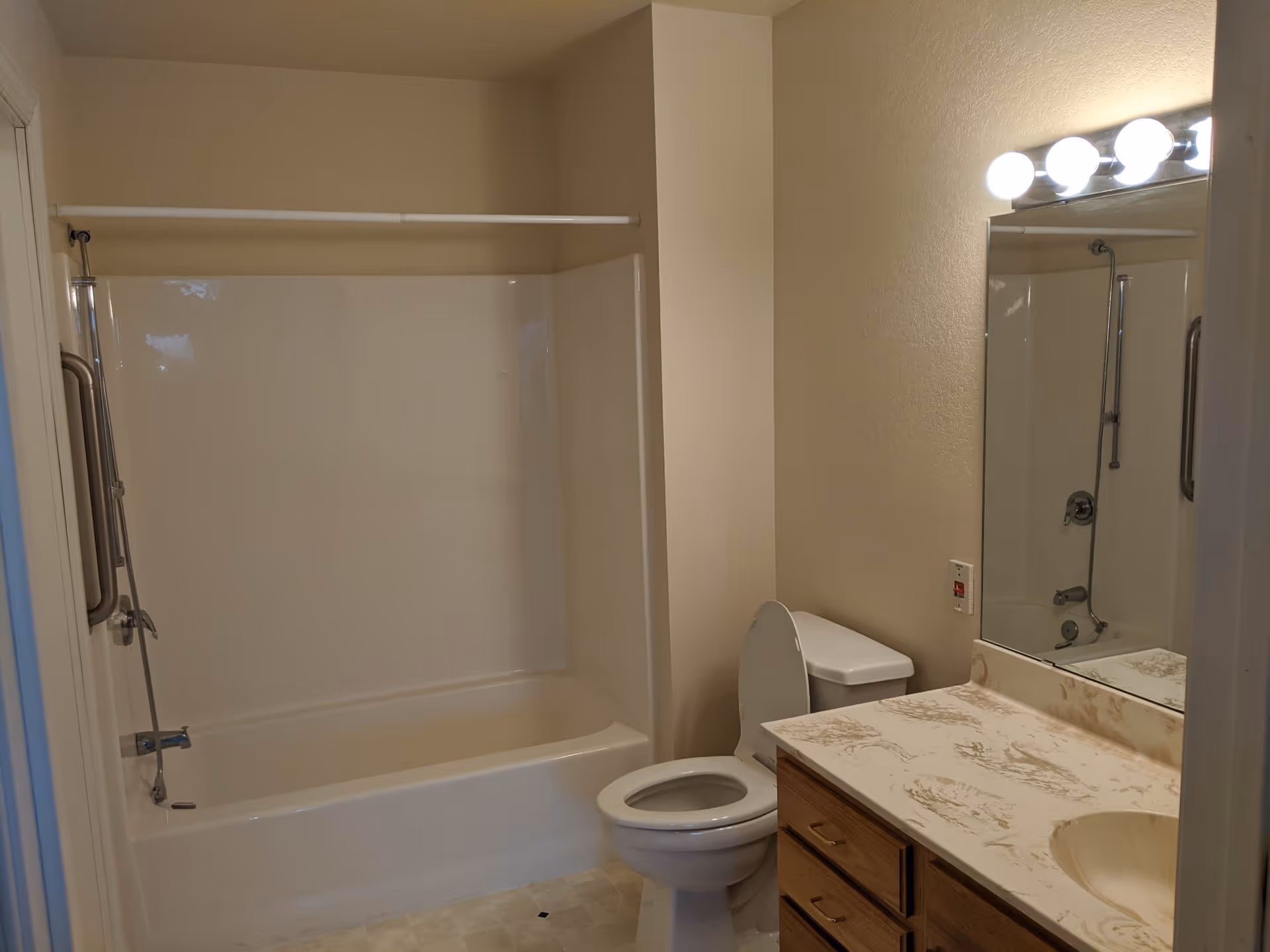 A bathroom with a white bathtub and shower combination, a toilet with the lid up, a countertop with a sink, wooden cabinets underneath, and a large mirror above the sink. There are four round light bulbs above the mirror providing illumination.
