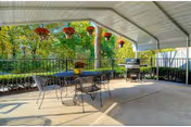 Covered outdoor patio area with a metal table and chairs, hanging flower pots, a barbecue grill, and a view of green trees and bushes in the background.