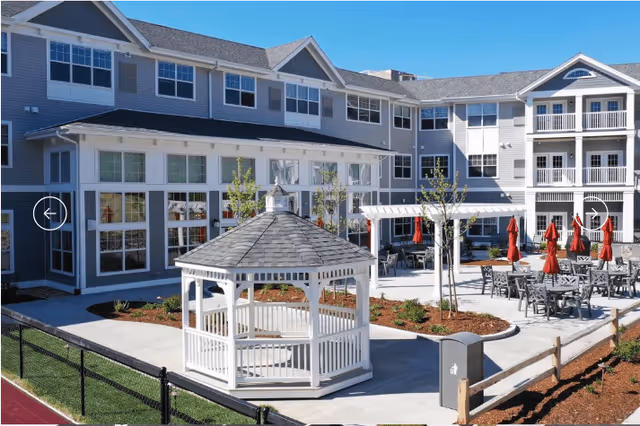 Outdoor courtyard area of a retirement community featuring a white gazebo, several tables with chairs and red umbrellas, young trees, and a multi-story building with many windows in the background under a clear blue sky.