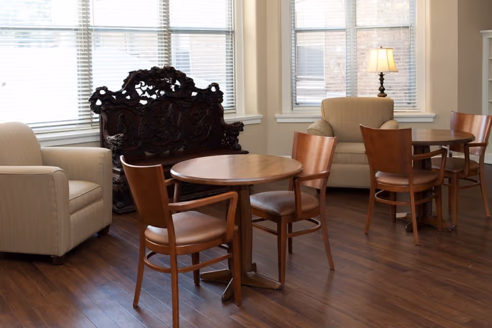 A bright common room with round wooden tables and chairs, upholstered armchairs, and a carved wooden bench by large windows.
