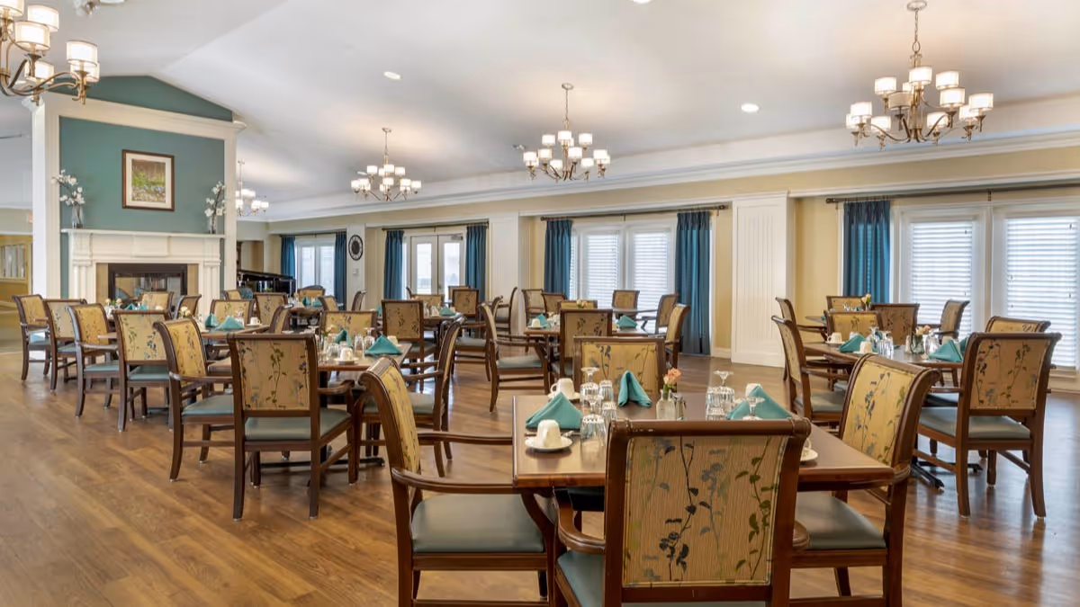 A spacious dining room with multiple wooden tables and chairs arranged neatly. Each table is set with teal napkins, glassware, and white cups. The room features hardwood flooring, large windows with blue curtains, and elegant chandeliers hanging from the ceiling. A fireplace with a framed picture above it is visible on the left side of the room.