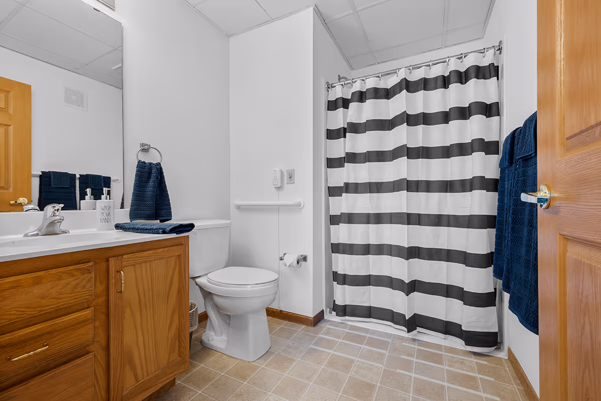 A clean bathroom with a wooden vanity and white sink on the left, a toilet in the center, and a shower with a black and white striped curtain on the right. Blue towels hang on the wall and on the vanity, and the floor is covered with beige tiles.