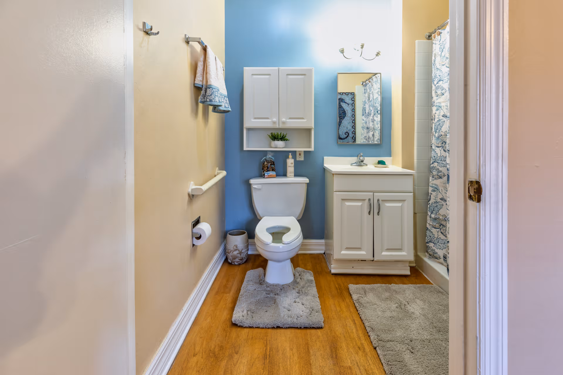 A clean bathroom with a toilet centered against a blue accent wall. Above the toilet is a white cabinet with a small plant and toiletries. To the right is a white vanity with a sink and mirror, and a shower with a blue and white patterned curtain. The floor is wood with two gray bath mats. Towels hang on a rack on the left wall.