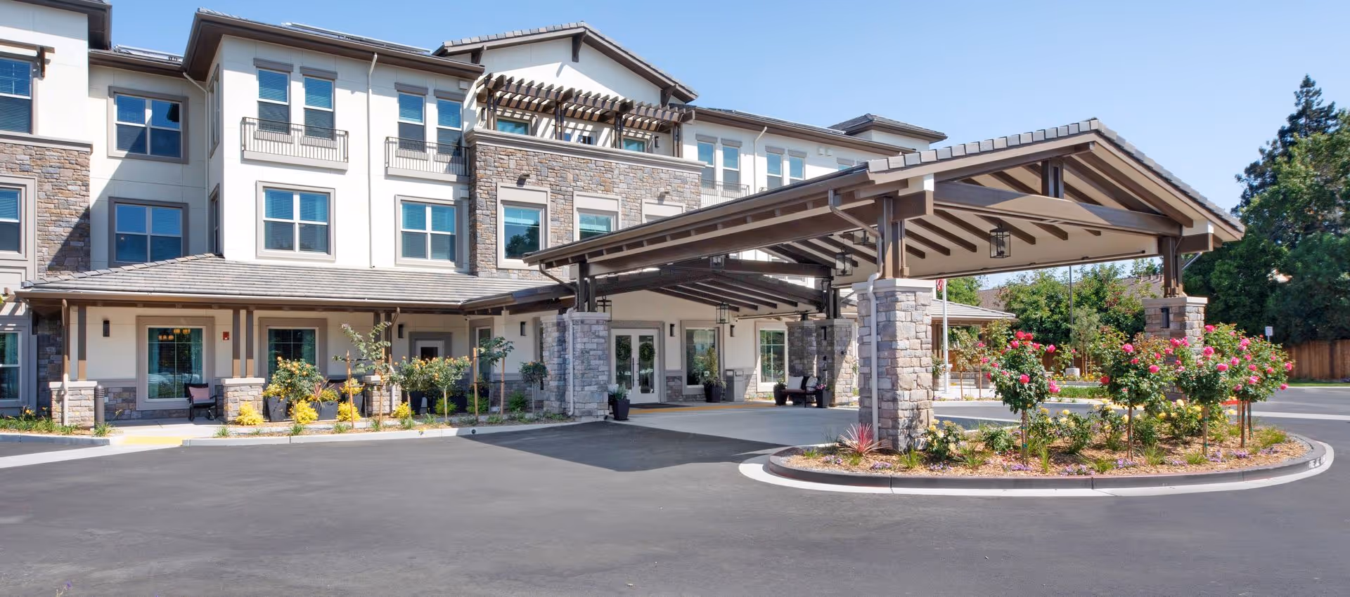 Front exterior of a senior living building with a covered porte-cochere, stone columns, and landscaped circular driveway.