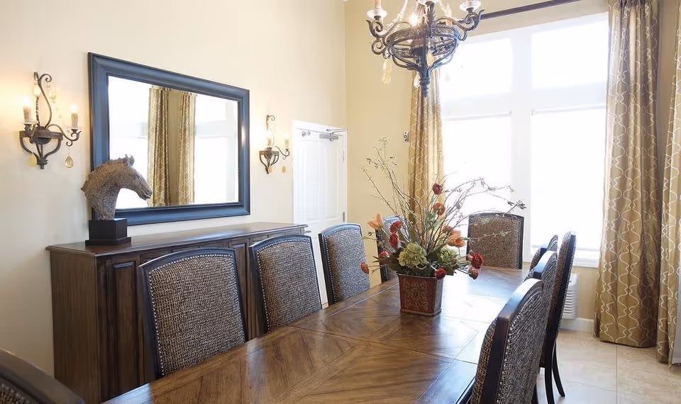 A bright dining room with a long wooden table surrounded by eight upholstered chairs. A floral centerpiece decorates the table. Behind the table is a wooden sideboard with a decorative horse head sculpture and a large mirror above it. Two wall sconces with candles flank the mirror. Large windows with patterned curtains allow natural light to fill the room.