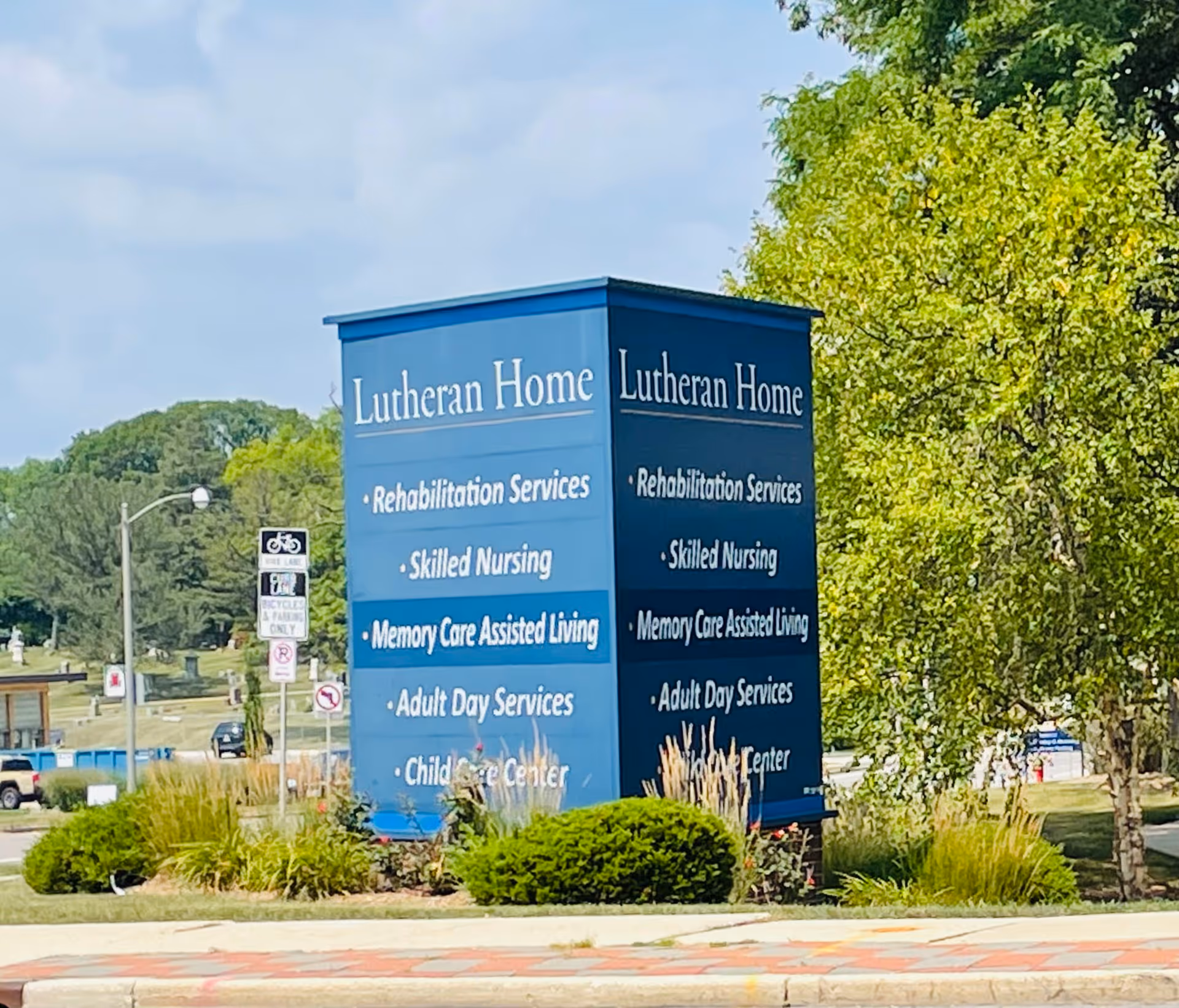 A large blue outdoor sign reading "Lutheran Home" listing services like rehabilitation, skilled nursing and memory care in a landscaped area with trees.