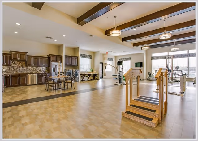 Spacious rehabilitation room with wooden handrails and steps for physical therapy, exercise equipment including stationary bikes and weight machines, a kitchen area with dark wood cabinets and stainless steel appliances, and a seating area with tables and chairs. Large windows allow natural light to fill the room.