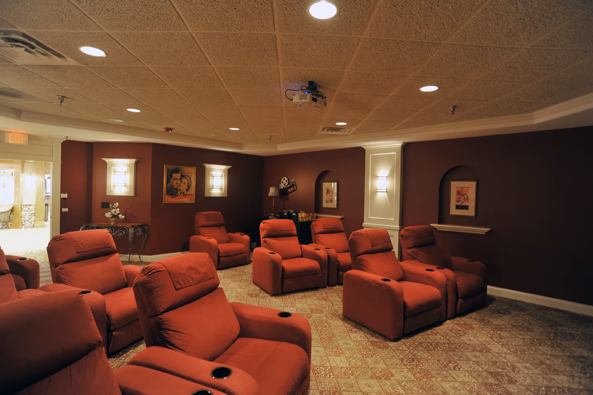 A cozy movie theater room with multiple red reclining chairs arranged in rows facing a projector mounted on the ceiling. The walls are painted dark red with framed vintage movie posters and soft wall sconces providing warm lighting. The carpet has a patterned design, and there is an exit door visible on the left side.