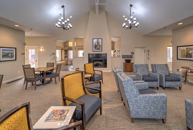 A spacious senior living common area with multiple seating arrangements including yellow and blue upholstered chairs, a fireplace in the center wall, framed artwork, and two chandeliers hanging from a vaulted ceiling.