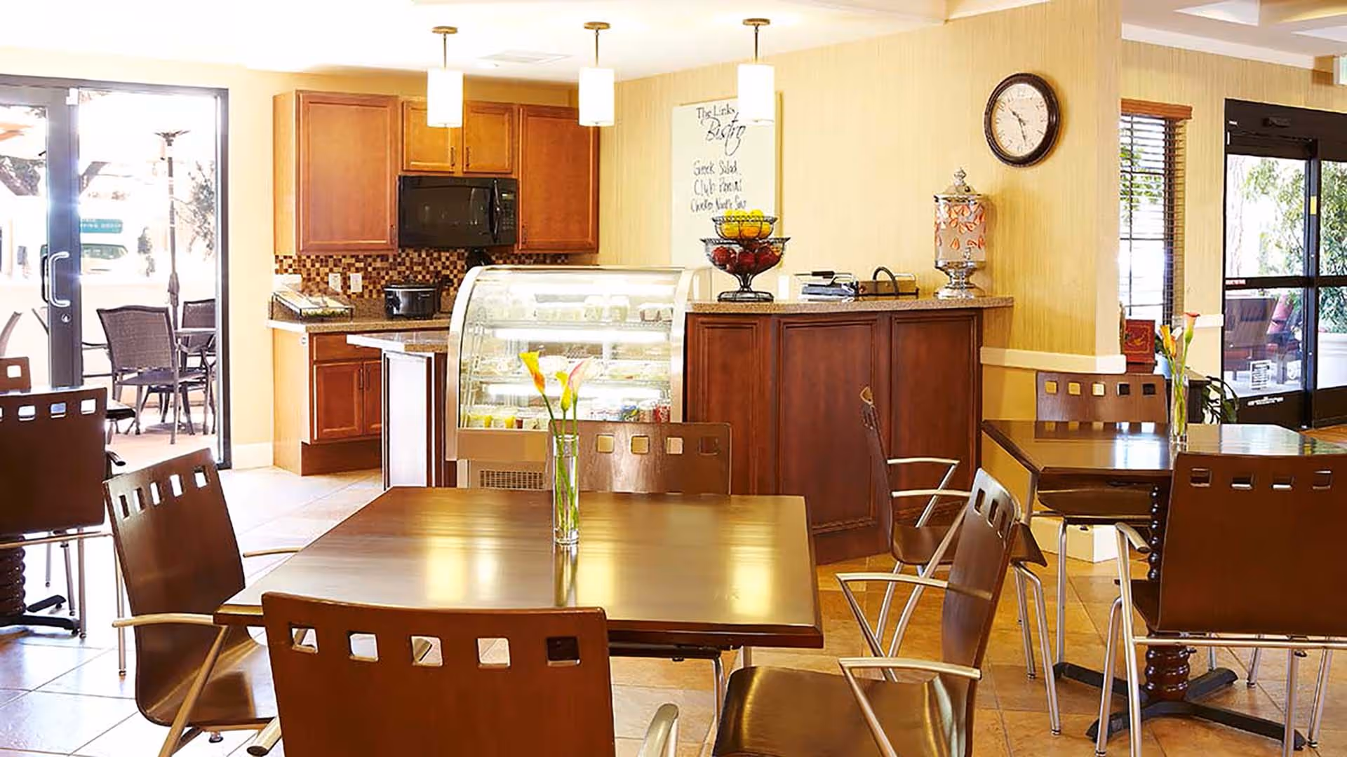A bright dining area with wooden tables and chairs, each table decorated with a vase holding flowers. In the background, there is a counter with a glass display case containing food items, wooden cabinets, a microwave, and a clock on the wall. Large glass doors lead to an outdoor seating area.