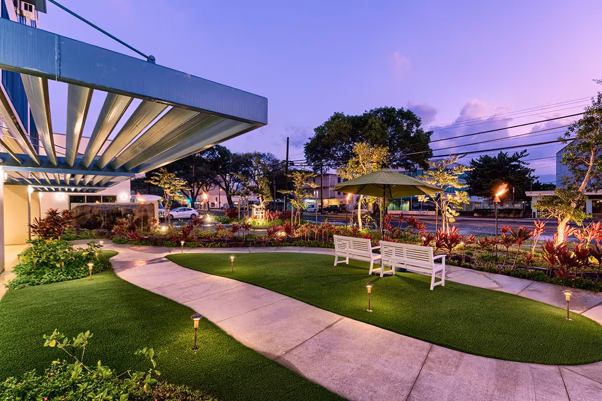 Evening view of a landscaped outdoor courtyard at Kalākaua Gardens with an illuminated walkway, benches, an umbrella, and a building canopy.