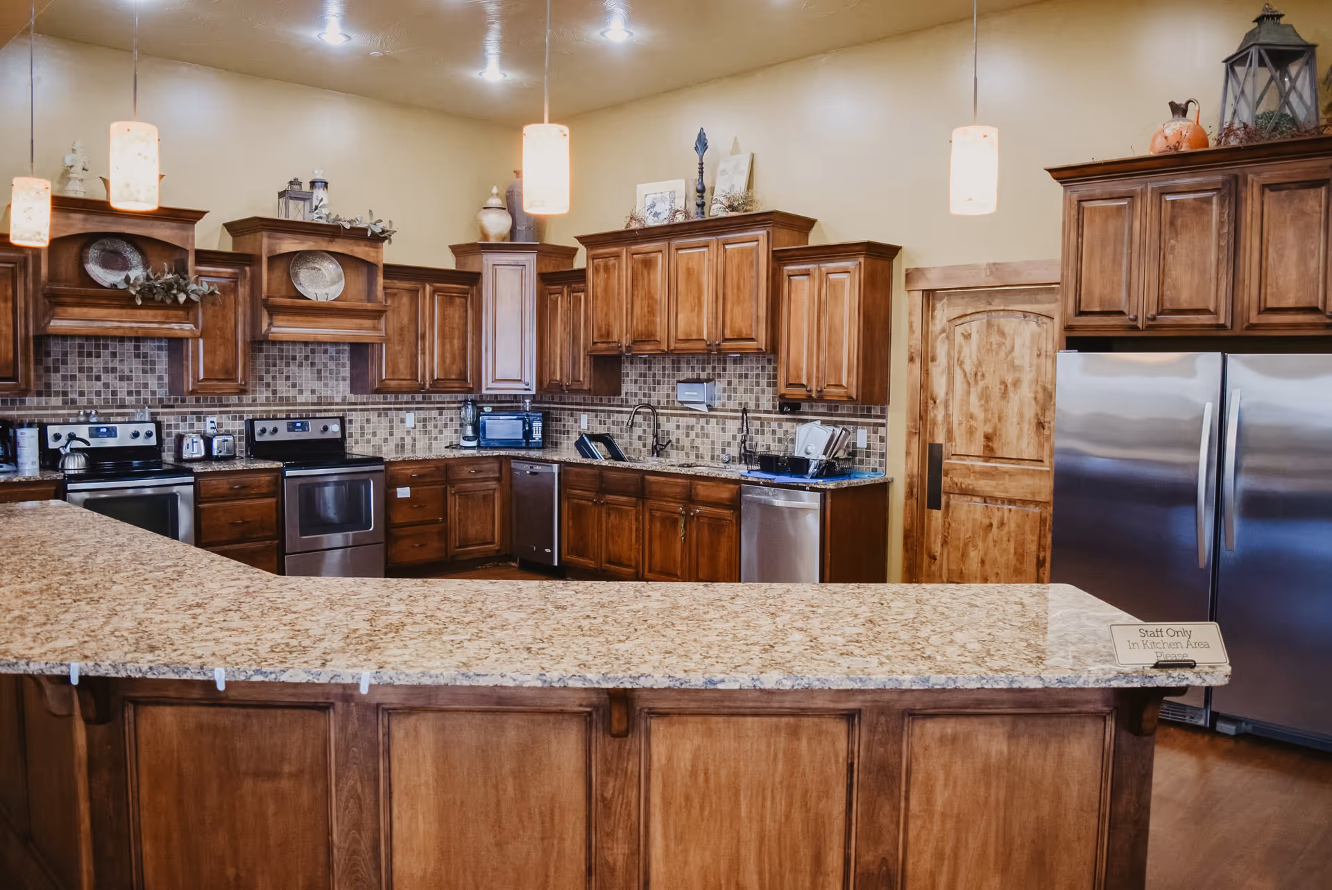 A spacious communal kitchen with wooden cabinetry, granite countertops, pendant lights, and stainless steel appliances.