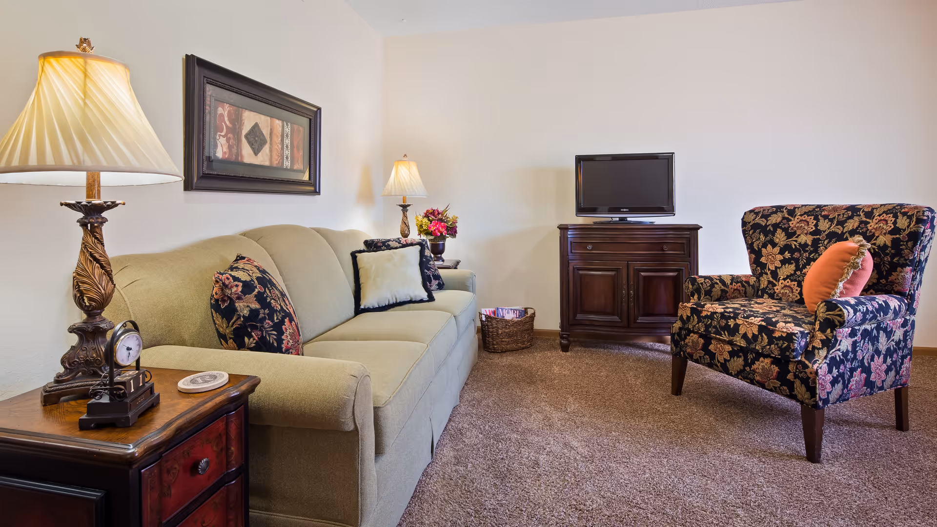 A cozy living room with a beige sofa adorned with floral and plain cushions, a floral patterned armchair with an orange pillow, a wooden side table with a decorative lamp and clock, a wooden TV stand with a flat screen TV, and a basket with magazines on a carpeted floor.