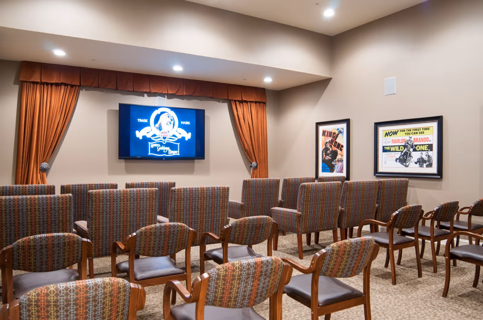 A small theater room with multiple rows of patterned upholstered chairs facing a wall-mounted flat screen TV. The room has beige walls, brown curtains framing the TV, and two framed movie posters on the right wall.