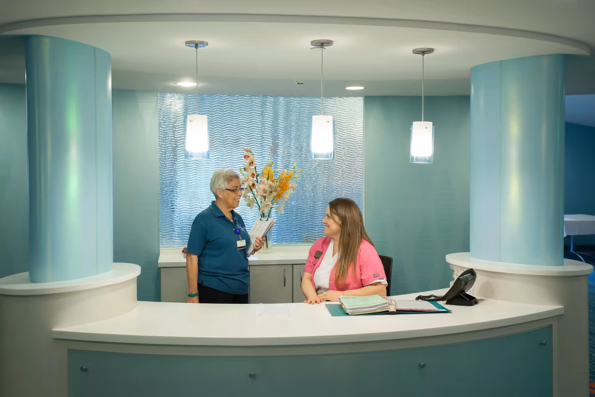 Two women at a curved reception desk in a well-lit interior space with blue walls and two large blue columns. One woman with gray hair and glasses is standing and holding a clipboard, while the other woman with long brown hair is seated behind the desk smiling. There are three pendant lights hanging from the ceiling and a vase with flowers on the counter behind them.