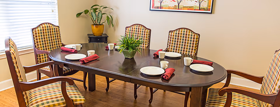 A dining room with a dark wooden oval table set with white plates, white mugs, and red cloth napkins. Six wooden chairs with plaid upholstery surround the table. A small green plant centerpiece is on the table. There is a window with blinds on the left and a framed artwork on the beige wall in the background.