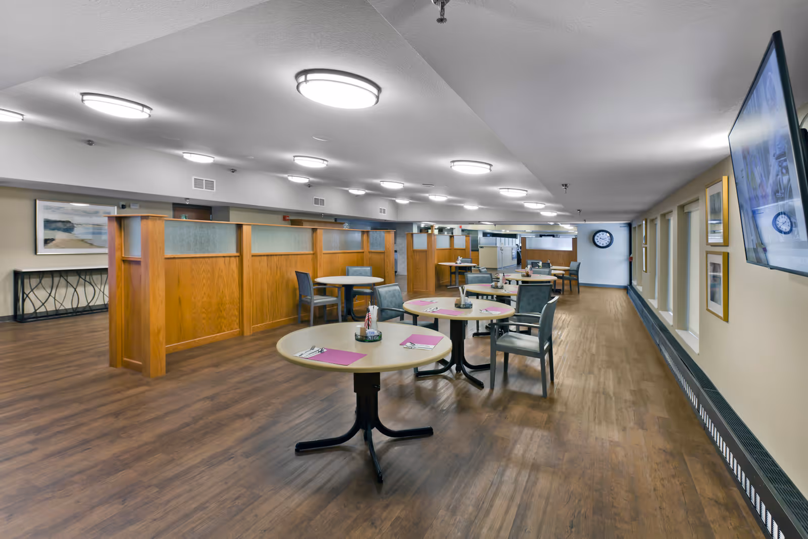 Interior view of a dining area in a senior living facility with round tables set with placemats, utensils, and small centerpieces. The room has wooden flooring, wooden partition panels, chairs around the tables, a wall-mounted TV, framed pictures on the wall, and ceiling lights.