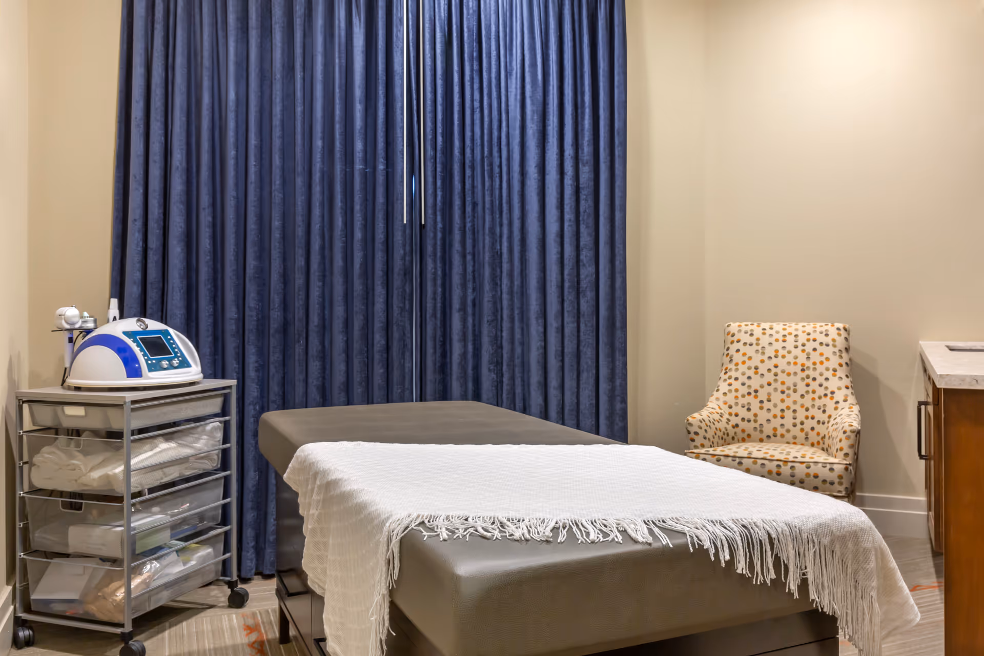 A therapy or treatment room with a padded treatment table covered with a white fringed blanket, a patterned armchair, a small metal cart with medical or therapy supplies, and dark blue curtains covering a window or wall.