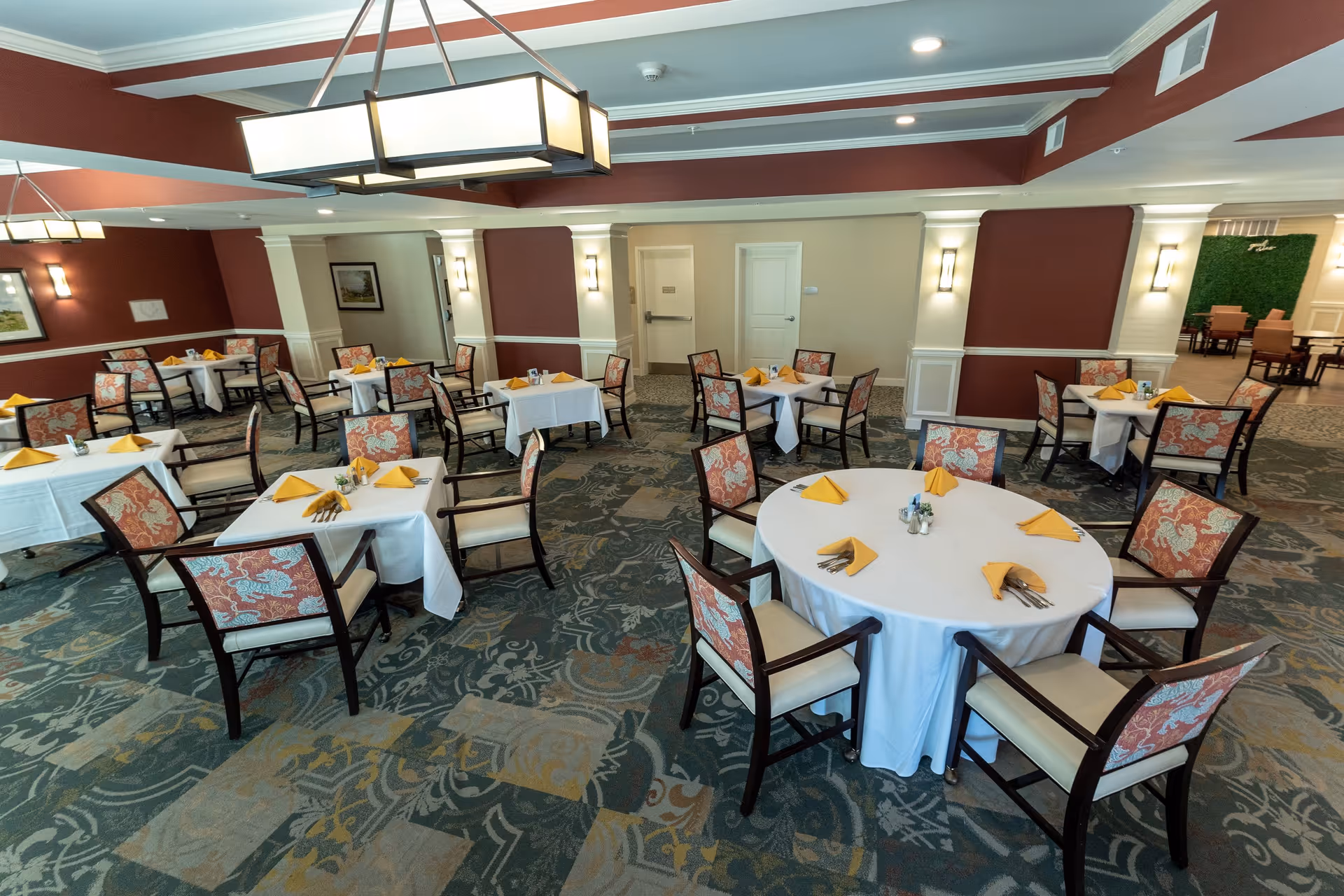 A spacious dining room with multiple tables covered in white tablecloths, each set with yellow folded napkins and silverware. The chairs have floral patterned upholstery. The room has maroon and beige walls with decorative lighting fixtures and a patterned carpet. There is an adjoining room visible in the background with more seating.