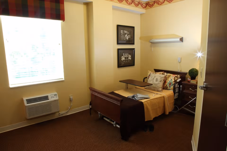 A small bedroom with a single bed covered in a yellow blanket and decorative pillows. There is a wooden nightstand with a small plant and a telephone. Two framed pictures hang on the wall above the bed. A window with a plaid valance lets in natural light, and an air conditioning unit is mounted below the window. The room has beige walls and brown carpet.
