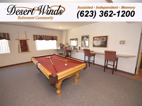 A recreation room with a red felt pool table in the center, two pool cues resting on it, and a single white cue ball. The room has beige walls, carpeted floor, and a window with a valance. Along one wall, there is a long counter with two high chairs and framed pictures above it. A rack holding pool cues is mounted on the wall near the window.