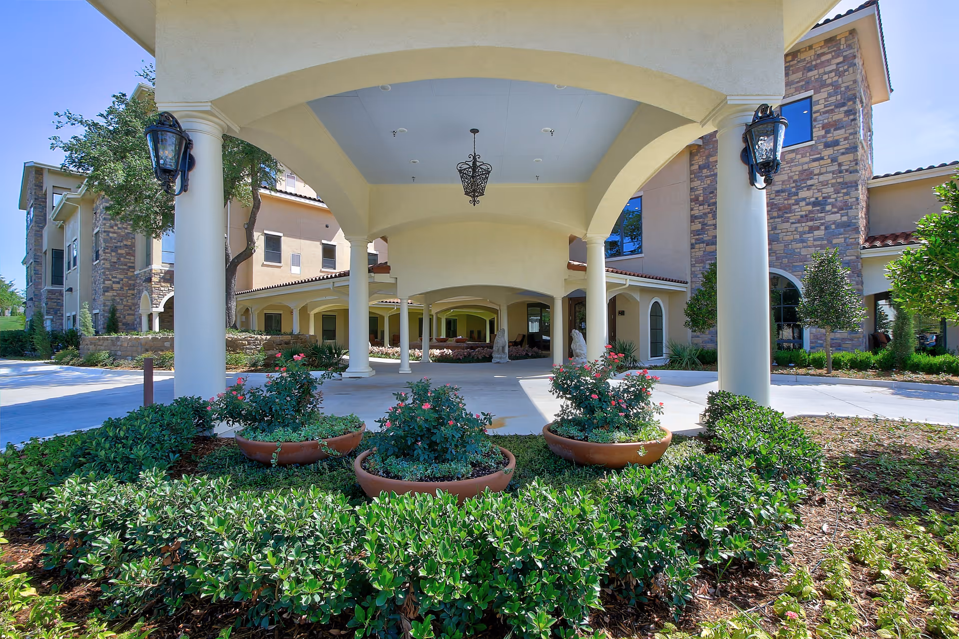 Covered driveway entrance of The Ridglea Senior Living facility with large white columns, hanging lantern-style lights, and landscaped flower beds with green shrubs and pink flowers in terracotta pots.