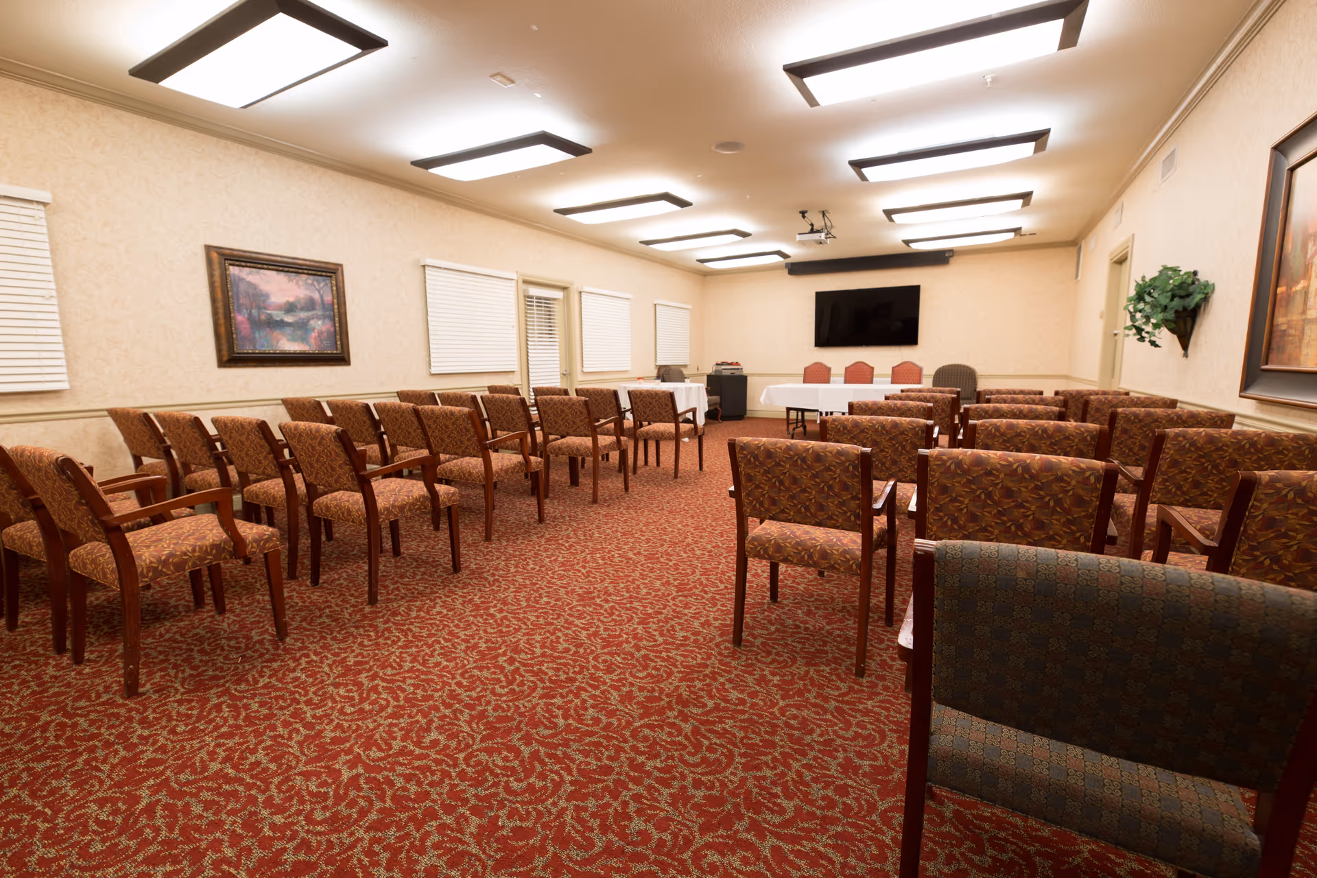 A meeting or conference room with rows of patterned chairs arranged facing a long table covered with white tablecloths. The room has beige walls, a red patterned carpet, several framed paintings, and a large flat-screen TV mounted on the far wall. Ceiling lights provide bright illumination.