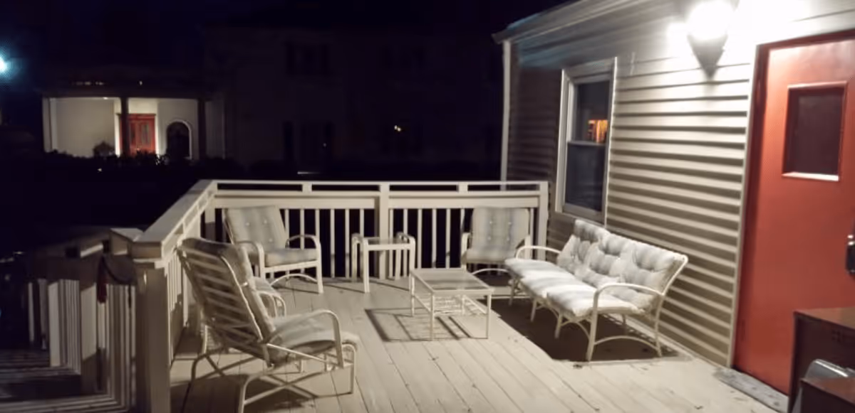 A nighttime view of an outdoor deck area with cushioned patio furniture including a sofa, three chairs, and two small tables. The deck is attached to a building with beige siding and a red door, illuminated by a wall-mounted light.