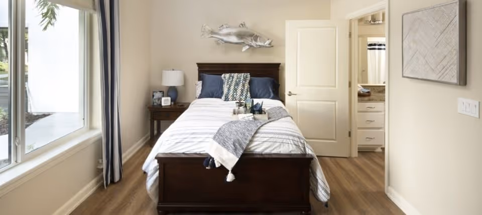 Sunlit bedroom with a centered wooden bed, nightstand, large window, and an open door leading to a bathroom.