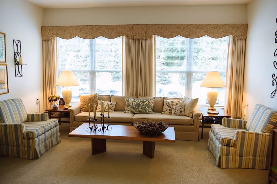 A cozy living room with a beige sofa adorned with decorative pillows, two striped armchairs, a wooden coffee table with a decorative bowl and candle holders, two side tables each with a lamp, and large windows with beige curtains letting in natural light.