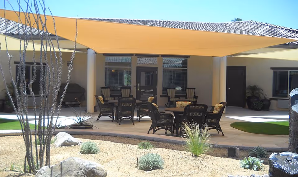 Outdoor patio area at Caleo Bay Assisted Living and Memory Care featuring a shaded seating area with multiple wicker chairs and tables, surrounded by desert landscaping with rocks, small plants, and a few taller desert plants.