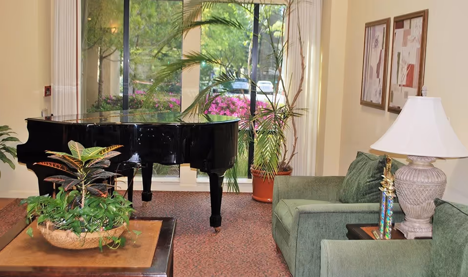 A cozy living room area with a black grand piano near large windows showing greenery outside. The room has green upholstered chairs, a side table with a lamp and a trophy, framed artwork on the wall, and a coffee table with a plant arrangement.