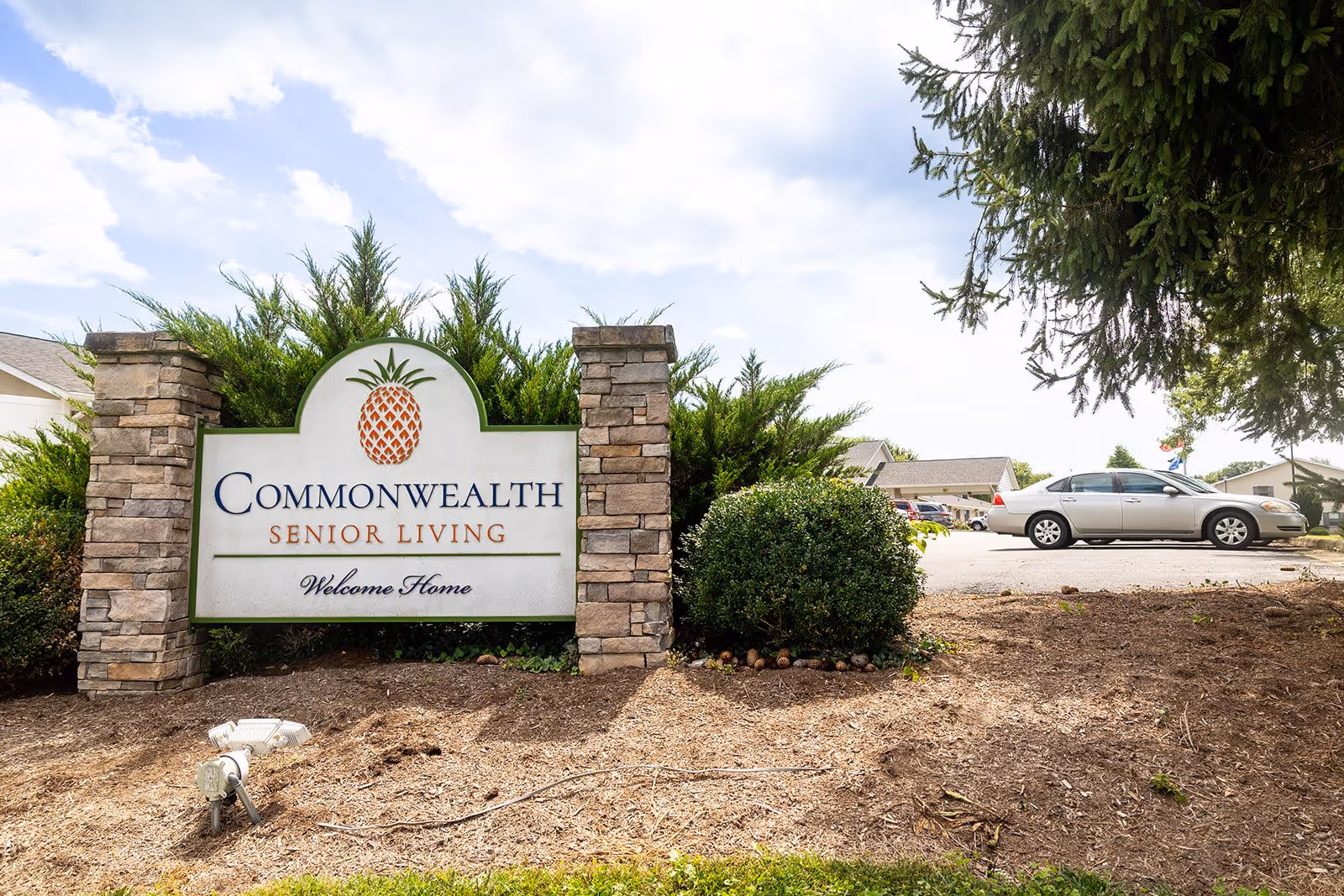 Outdoor view of the entrance sign for Commonwealth Senior Living, featuring a stone frame and a pineapple logo, with a parking lot and cars in the background under a partly cloudy sky.
