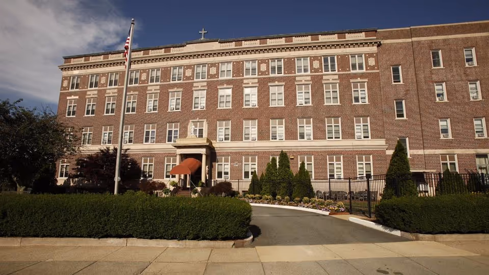 Front exterior of a large multi-story brick nursing facility with an American flag, entrance awning, and landscaped hedges.