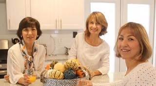 Three women smiling and standing around a kitchen island with a bowl of decorative gourds.