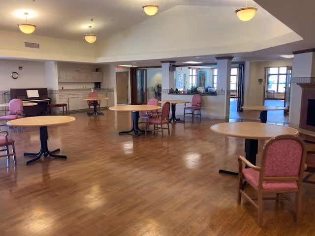 Open senior facility common room with round tables and pink upholstered chairs on wood flooring, a piano and kitchenette in the background.