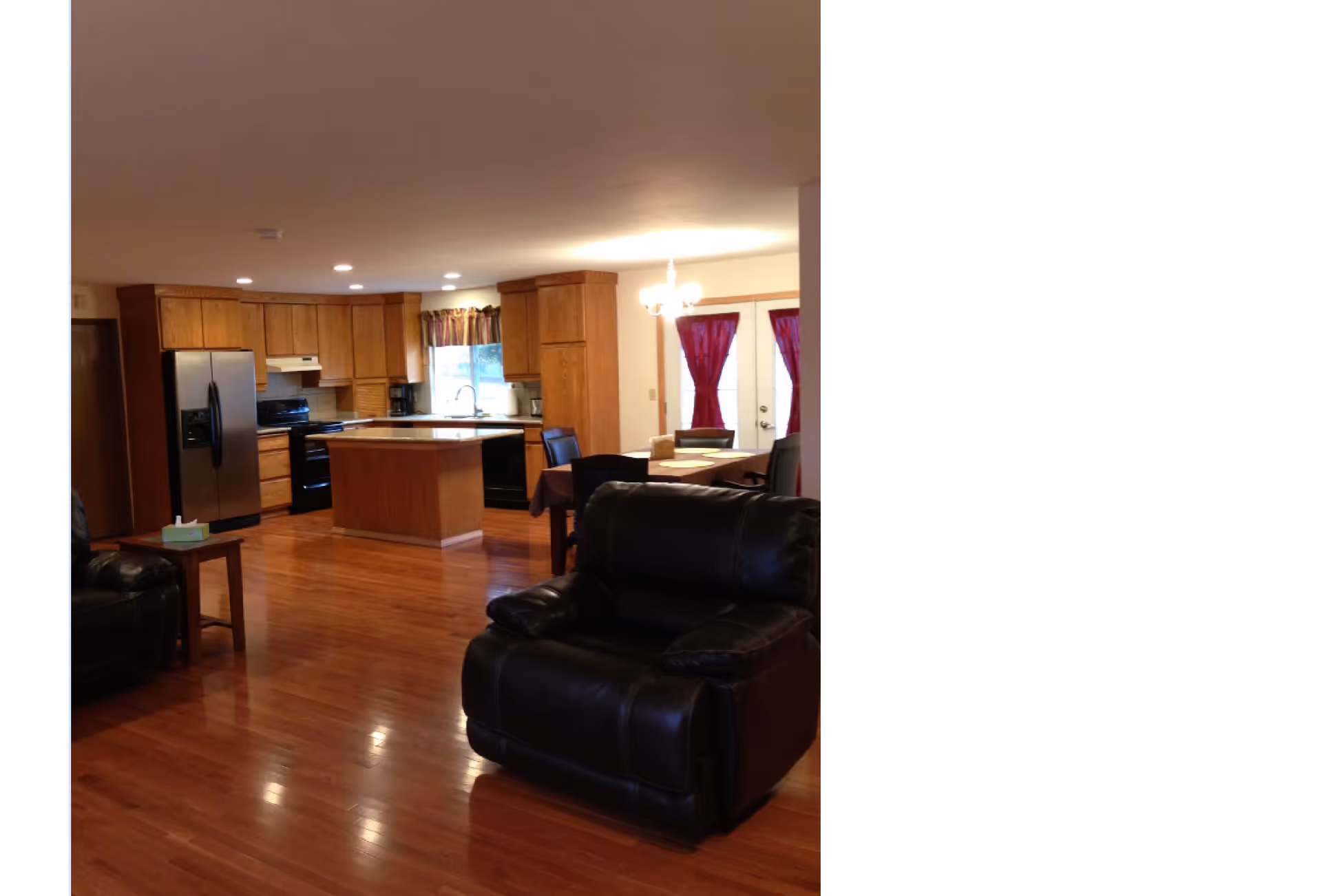 Open interior space showing a kitchen with wooden cabinets, a central island, stainless steel refrigerator, and black appliances. Adjacent to the kitchen is a dining area with a table and chairs near glass doors with red curtains. In the foreground, there is a black leather armchair and a small wooden side table with a tissue box on it. The floor is polished wood and the ceiling has recessed lighting.